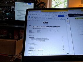 Das Foto zeigt einen Computerbildschirm, auf welchen die erste Seite des Protokoll der StuRa-Sitzung zu sehen ist. Dahinter ist auf einen zweiten Bildschirm die Videokonferenz der Sitzung zu sehen.