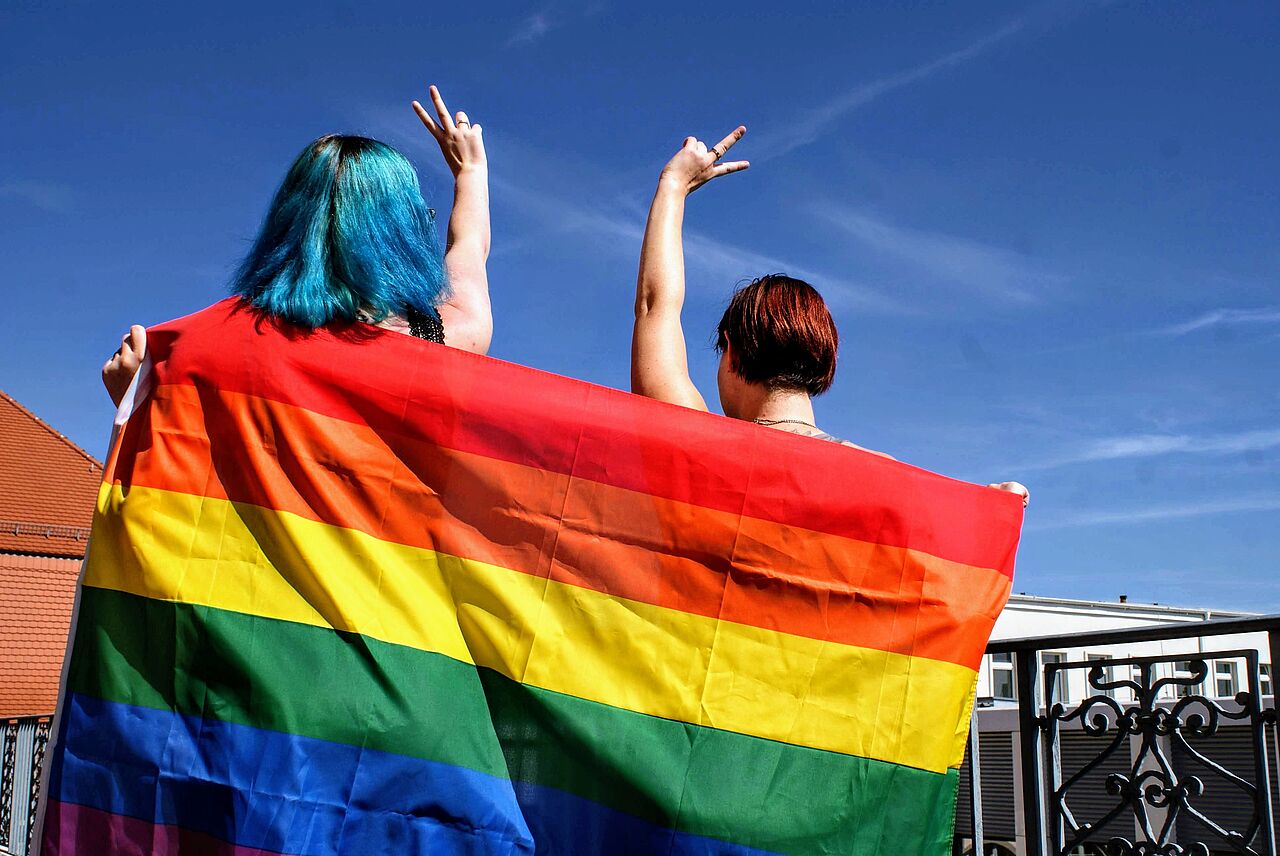Marion und Maria strecken die bunte Pride-Flagge und ihre Hände mit dem „Peace”-Zeichen Richtung Himmel